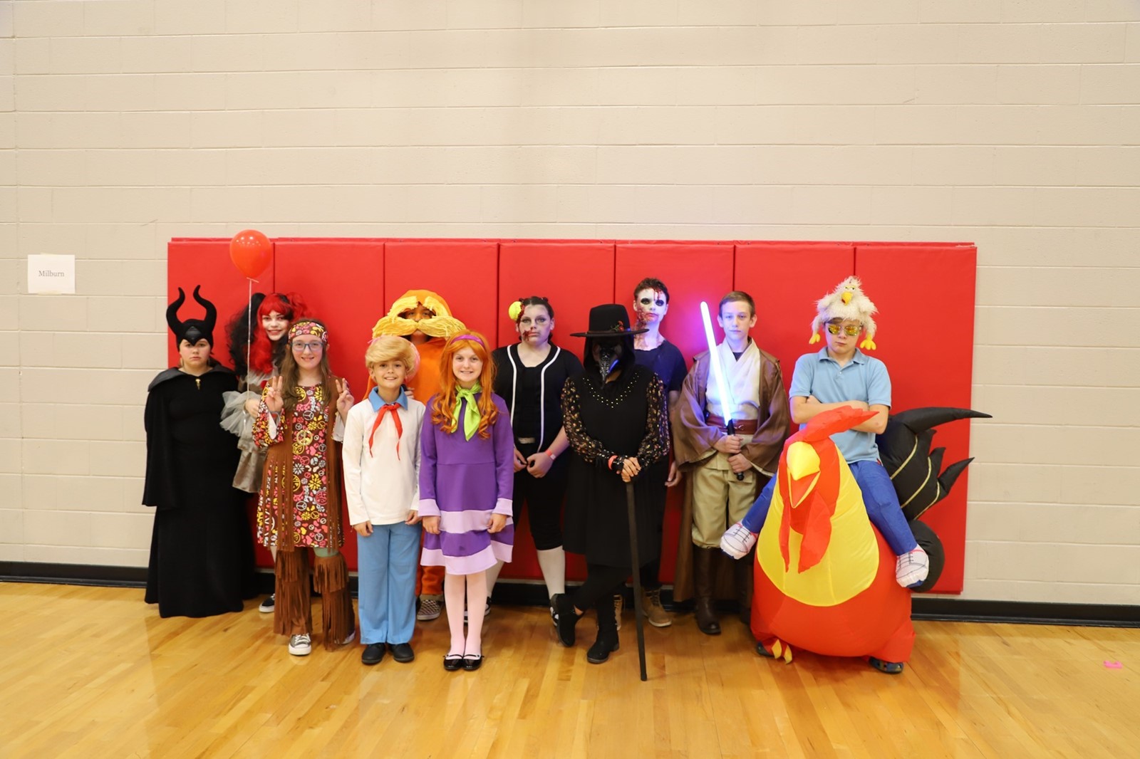 A group of students stand in front of a gym mat dressed in Halloween costumes. 