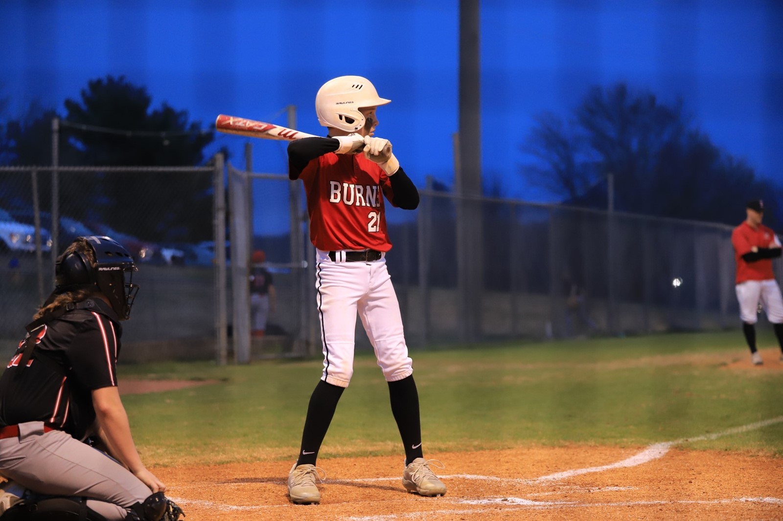 Child in red baseball outfit stands at bat on a field in the evening. 